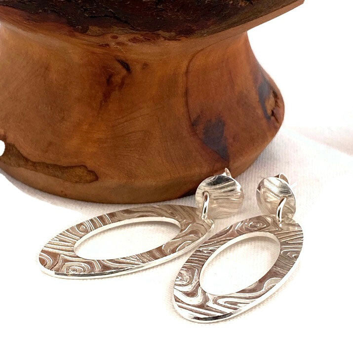 Engraved Oval earrings beside wood, showing swirling engraved detail and round stud tops